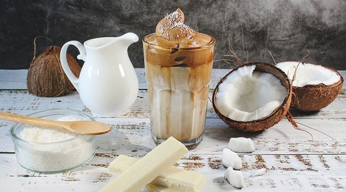 iced coffee on a table with coconuts, a glass sugar container, pitcher of milk, and two pieces of white chocolate bars