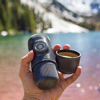 Person holding the Wacaco Nanopresso Espresso Maker with one hand in front of the lake and trees 