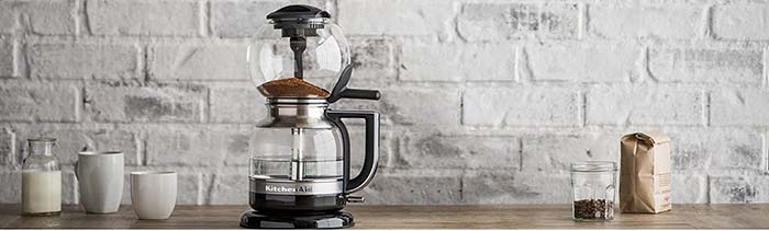 KitchenAid Siphon Coffee Maker on a wooden kitchen counter beside two white ceramic mugs, a glass of milk, & coffee beans