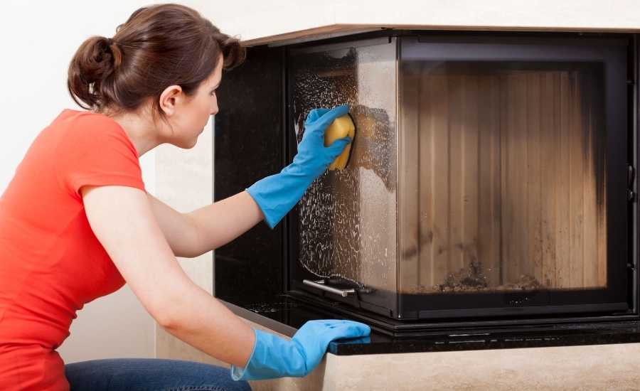 A woman in an orange shirt and blue rubber gloves, wiping the glass of the fireplace with a yellow sponge