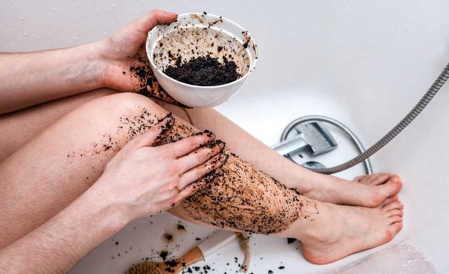 A woman sitting in a tub & scrubbing her right leg with coffee grounds made into a paste