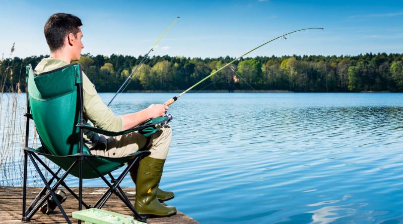 A man wearing a light green shirt, khaki pants, and dark green boots sits on a green camping chair and is lake fishing